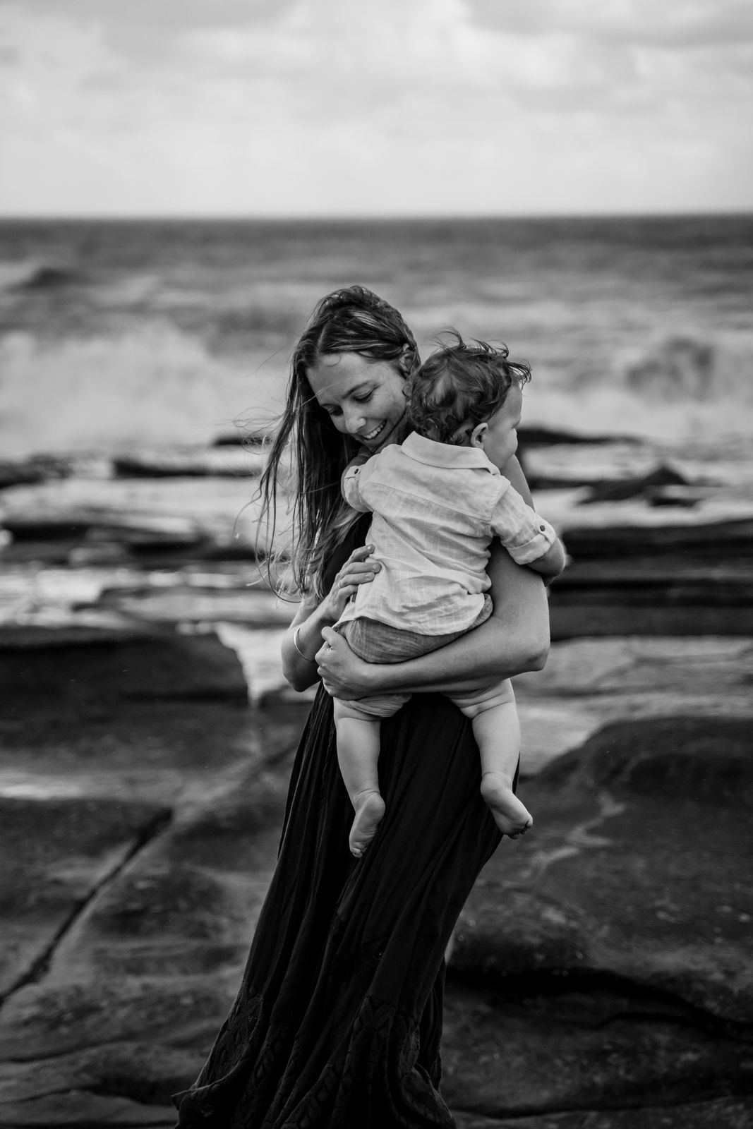 Black and white photo of a mum holding her toddler on the rocks by the sea