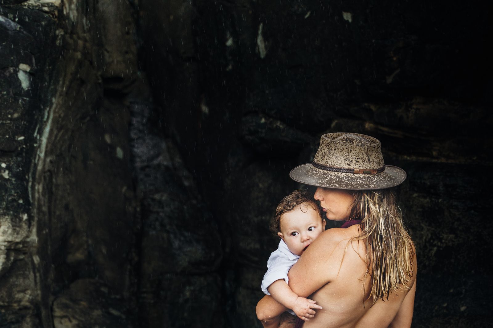 Mum in a sun hat holding her baby skin to skin against dark rocks