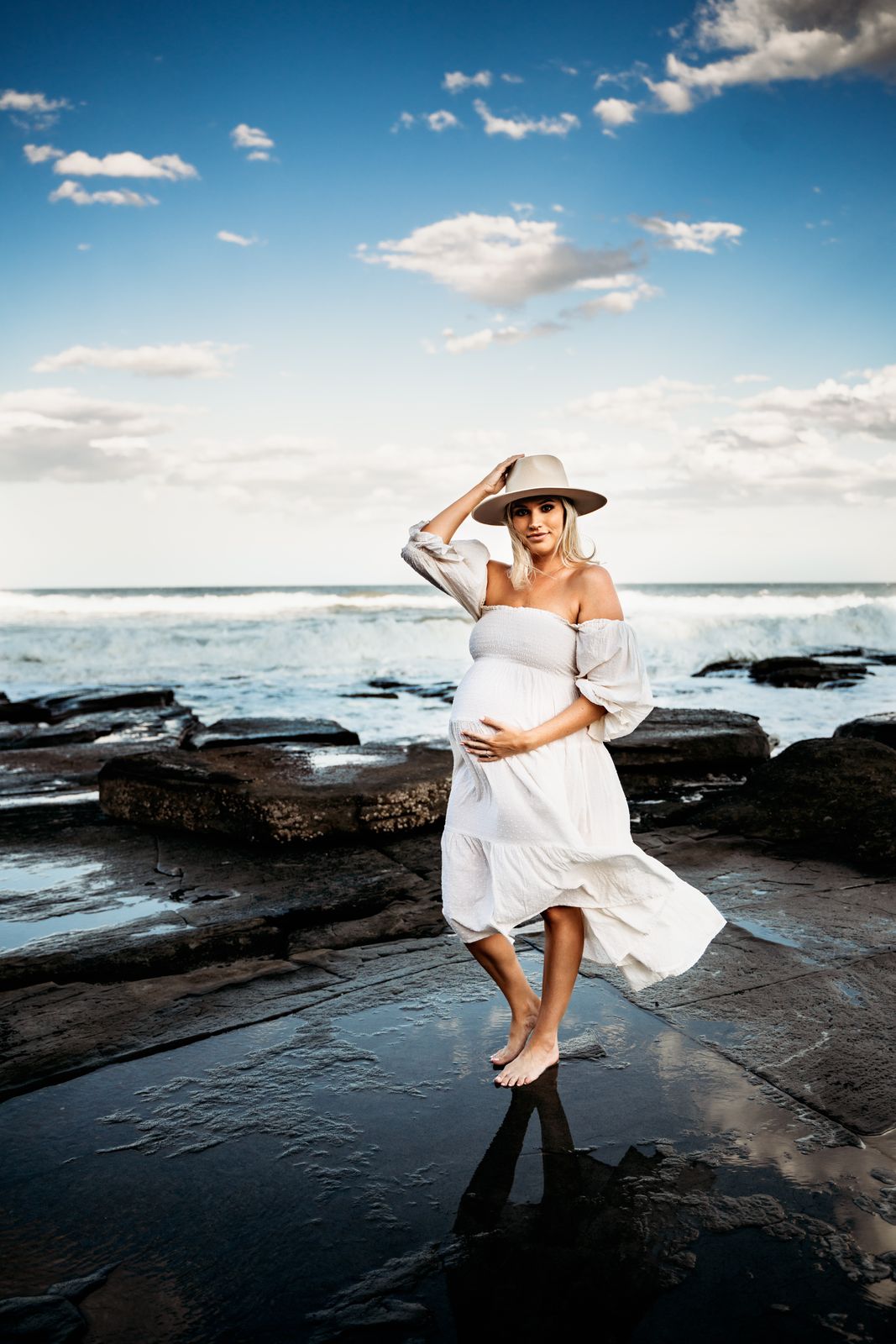 Mum-to-be in a white dress and hat on the rock shelf under a blue sky