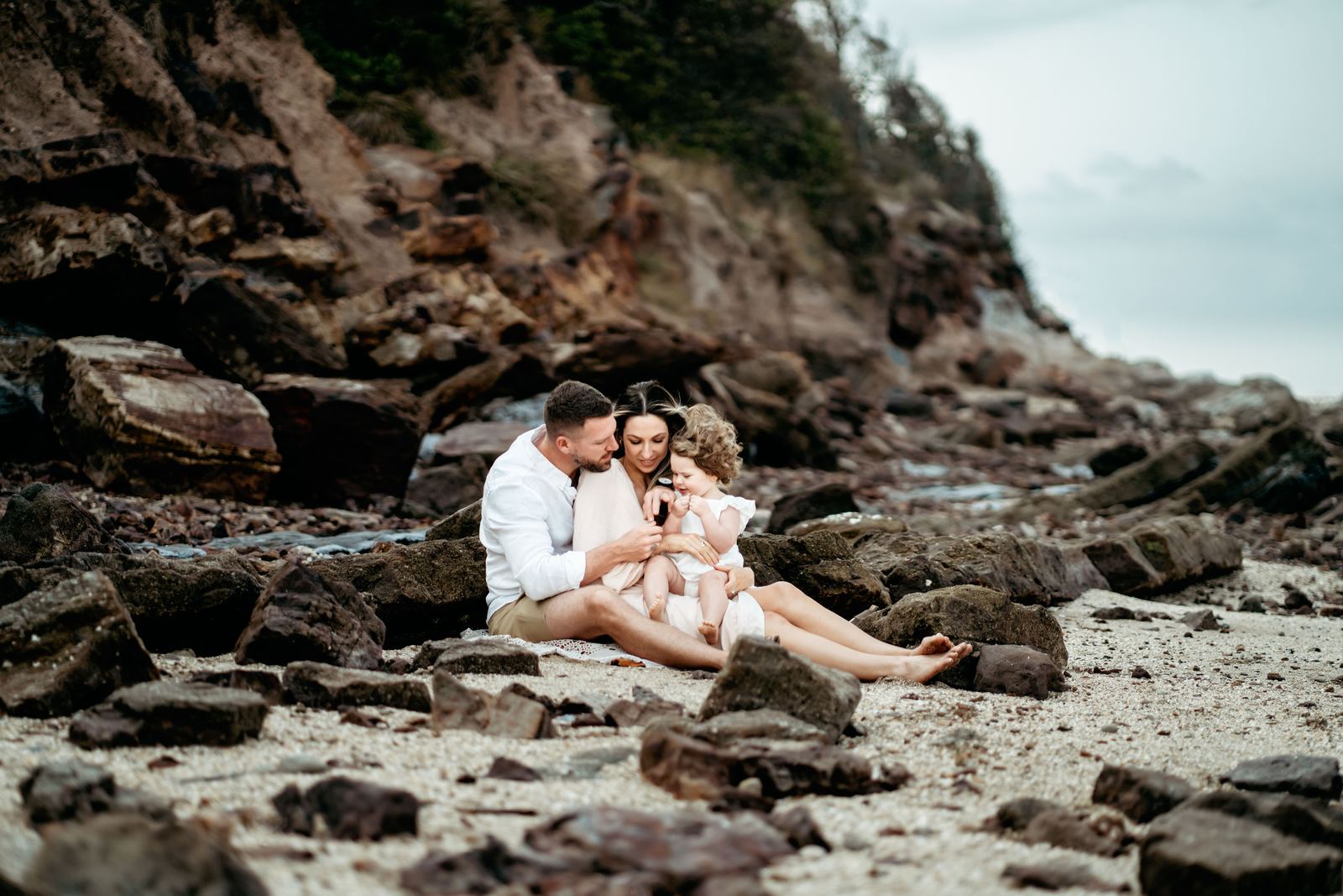 Young family sitting close together on a rocky beach under a cloudy sky